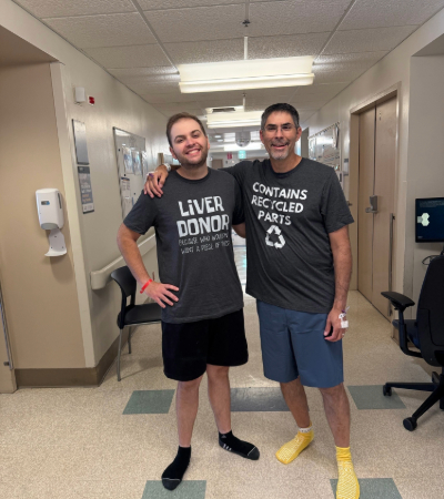 Two people in a hospital hallway wearing matching gray shirts, one labeled &ldquo;Liver Donor&rdquo; and the other &ldquo;Contains Recycled Parts.&rdquo;