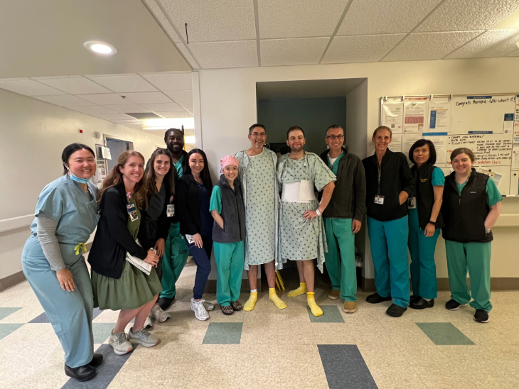 A group of hospital staff in scrubs and two patients in gowns stand together in a hallway, posing for a photo.