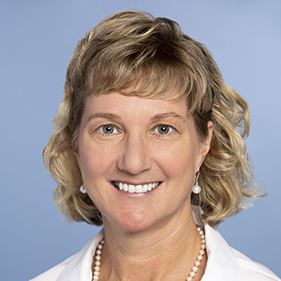 Headshot of smiling blonde woman with pearl earrings, a pearl necklace and the white collar of a doctors coat.