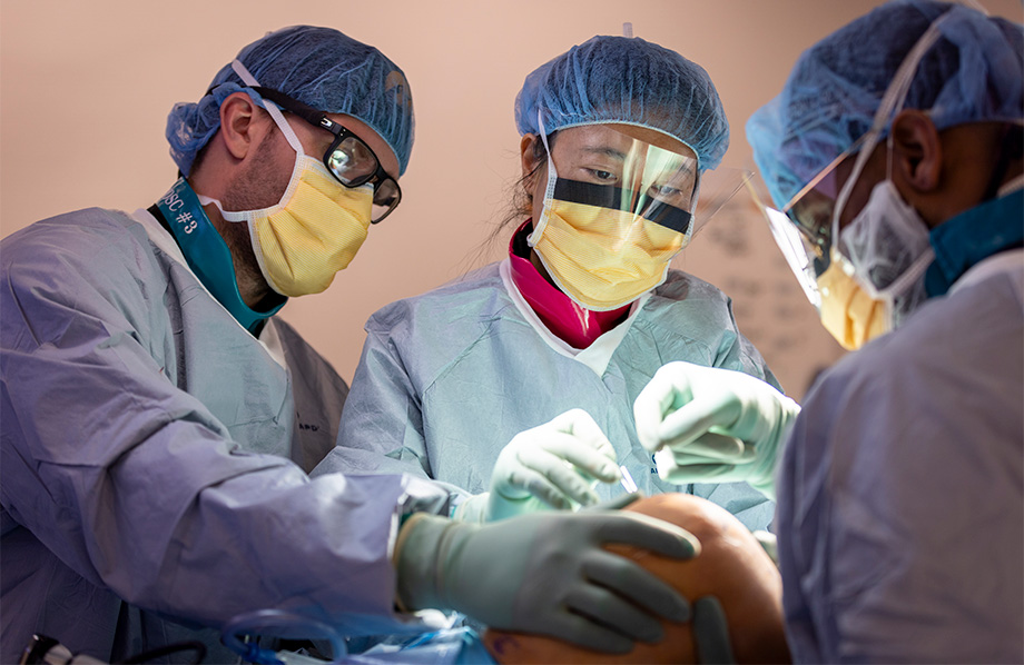 Three surgeons in medical scrubs gather around a patient&rsquo;s knee as they perform a surgery in an operating room. 