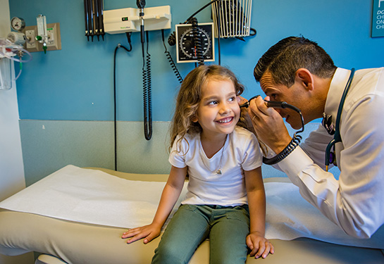 Doctor looks in girl’s ear while she smiles on patient table.  