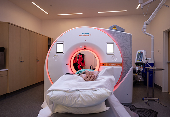 Large white cylindrical CT scanner with orange light and patient’s hands crossed in the foreground.
