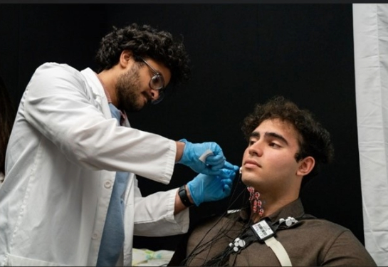 Harsha Gowda stands wearing a white coat placing electrodes on Ramie Wakeb who is sitting in a chair.