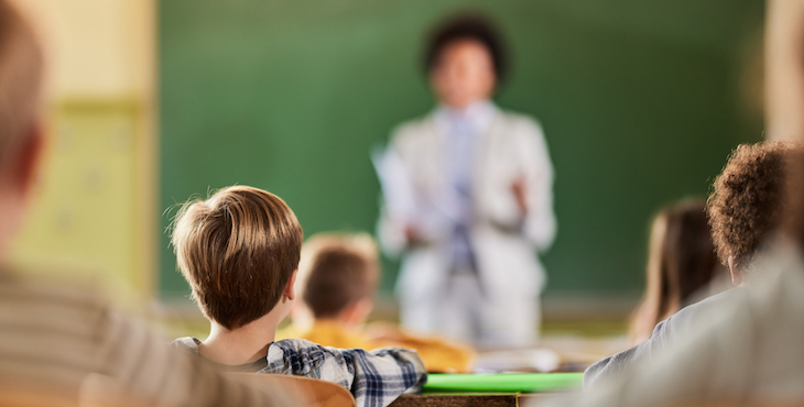 The back of students&rsquo; heads are shown sitting in rows in a classroom with a teacher and green blackboard out of focus in the front of the room.