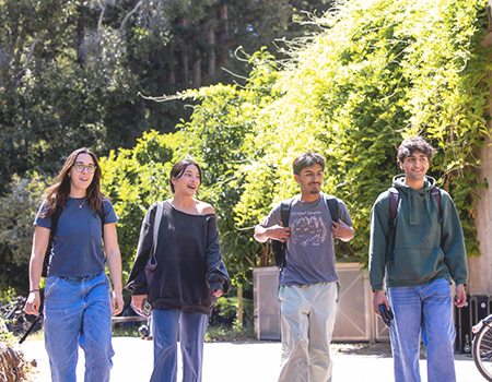 A group of four students wearing backpacks walk side by side across the college campus at UC Santa Cruz.