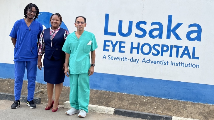 Two men and one woman stand in front of a white sign with blue lettering that says &ldquo;Lusaka Eye Hospital.&rdquo;