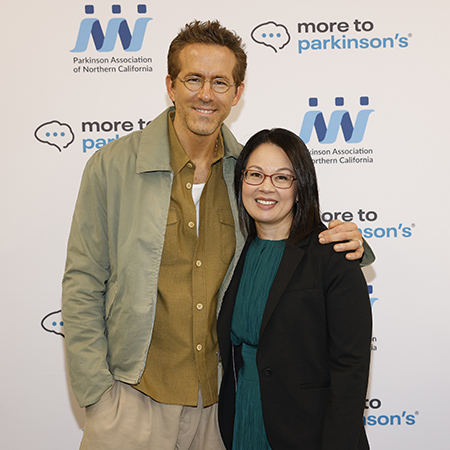 Ryan Reynolds is in light green blazer, olive shirt and khaki trousers. Norika Malhado-Chang is in green dress and black jacket. Both pausing for camera.