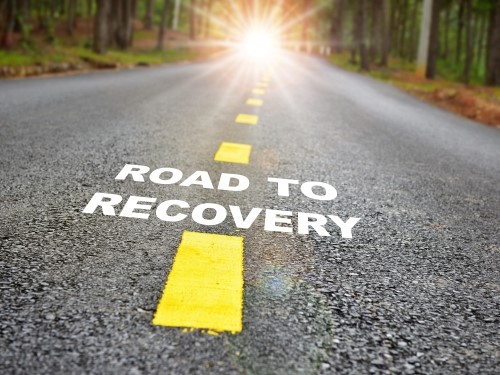A paved road with yellow lines in a forest with the words ROAD TO RECOVERY.
