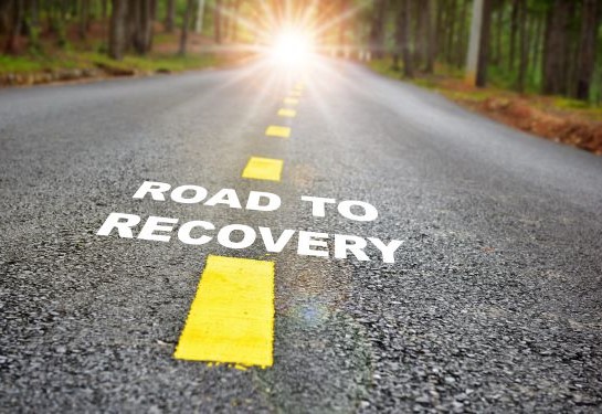 A paved road with yellow lines in a forest with the words ROAD TO RECOVERY.