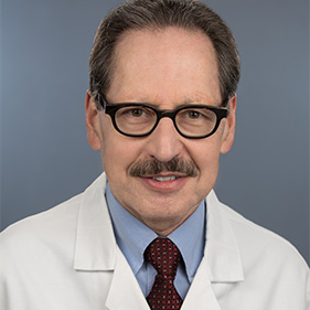 A man with brown hair and wearing glasses and a white physician’s coat, smiles