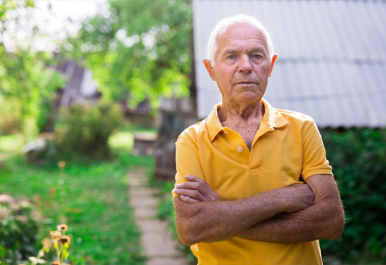 An older man stands outdoors with his arms crossed, looking directly at the camera in a green, rural setting.
