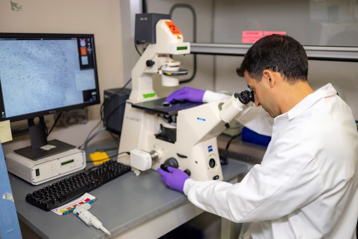 Cameron Sadegh is wearing his lab coat and purple gloves. He is looking at a tissue sample through an electronic microscope.