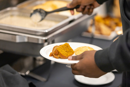 A person holds a white plate with two pieces of bread or cake near a buffet-style setup with trays of food in the background.