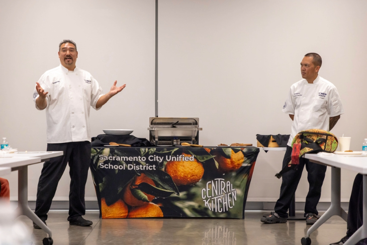 Two chefs stand at the front of a classroom-style setting, speaking to an audience.