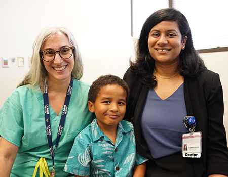 A boy sits between his surgeon in scrubs and his neurologist in business clothes.  All are smiling.
