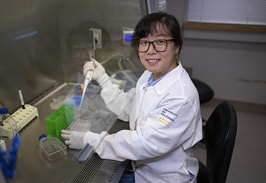 Woman with dark brown hair wearing a white lab coat and holding an instrument in a laboratory fume hood