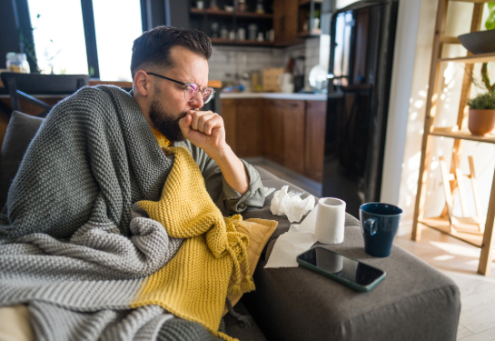 Sick man on sofa coughing, wrapped in blanket, surrounded by tissues and toilet paper, feeling unwell.