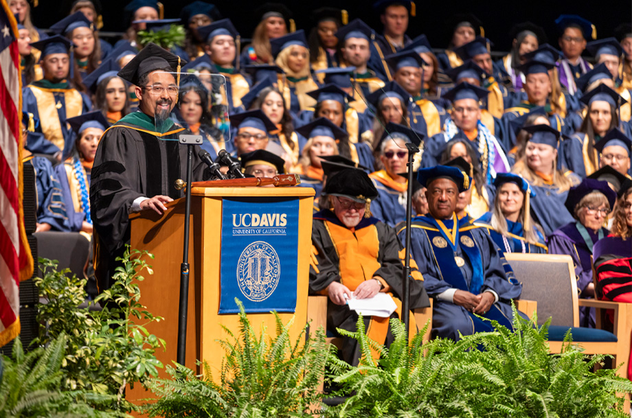 Keynote speaker speaks to audience from podium while faculty and graduates sit on risers on stage