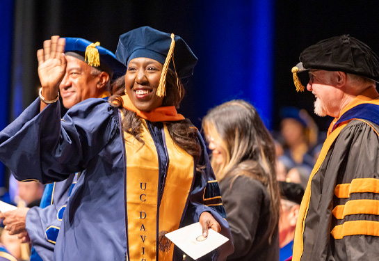 Woman in cap and gown waving to crowd while walking across stage