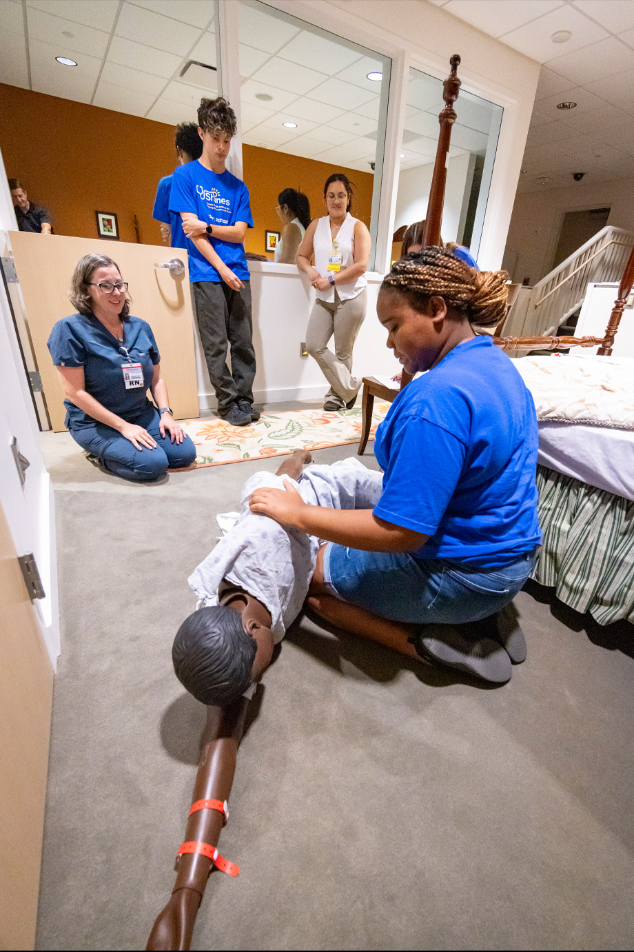 Student wearing SHINES blue shirt practices rolling a mannikin to its side in front of a bed as another student and nurse watch