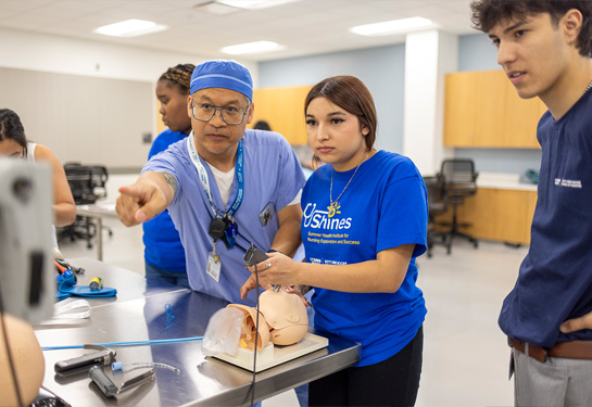 Nurse in scrubs points at monitor while student in blue SHINES shirt holds medical device in the mouth of mannikin on table