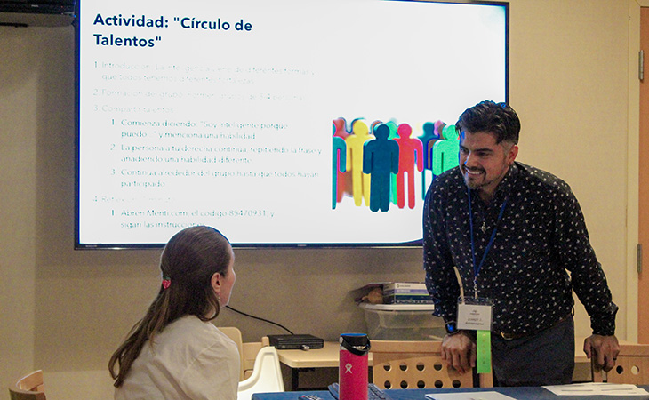 A woman, seated, talks to a man, standing, in front of a whiteboard containing a presentation in Spanish.