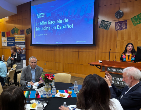 People sit around tables in a room where a projection screen shows a slide that states, &ldquo;La Mini Escuela de Medicina en Espa&ntilde;ol&rdquo;