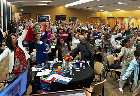 Dozens of men and women in a large conference room stand while swaying their arms during a group exercise