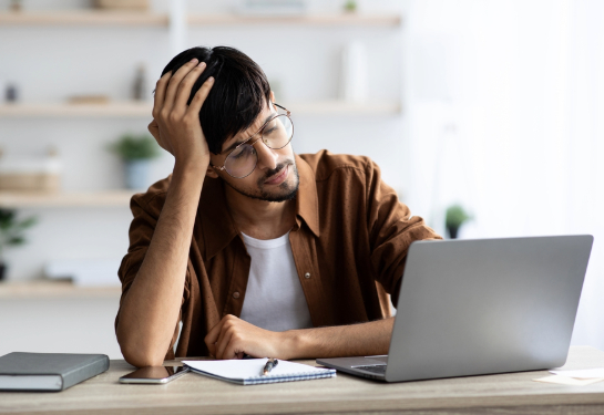 Exhausted man sitting in front of laptop at office, touching his head.