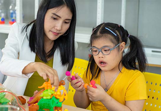 A medical provider in a white coat sits at a table with a child playing with colorful plastic toys
