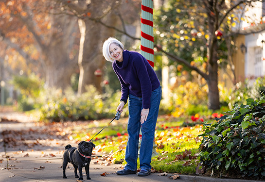 Woman with short grey hair, wearing purple sweater, leans over pug dog on street corner.