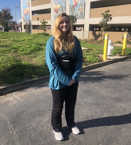 Tiffany Balts is standing in front of a parking structure with a flower mural. She is smiling and wearing a blue shirt and black pants.