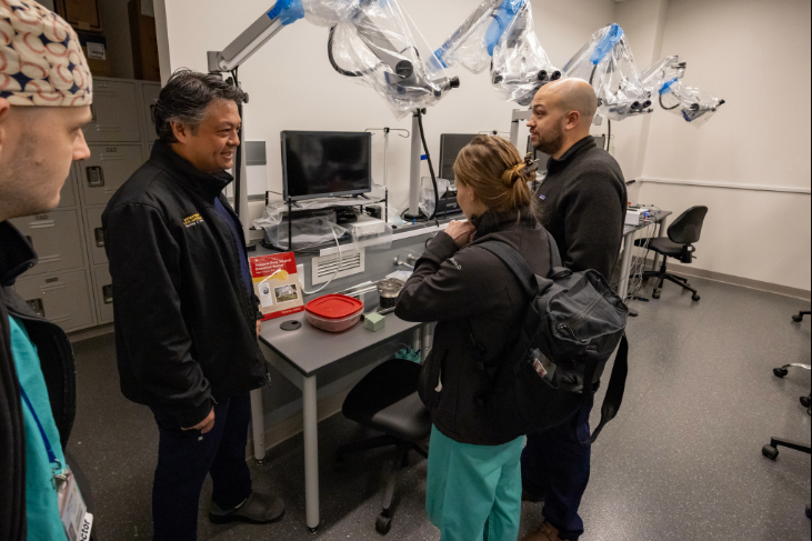 Three people standing near a lab workstation with monitors, microscopes and medical equipment, observing the setup.