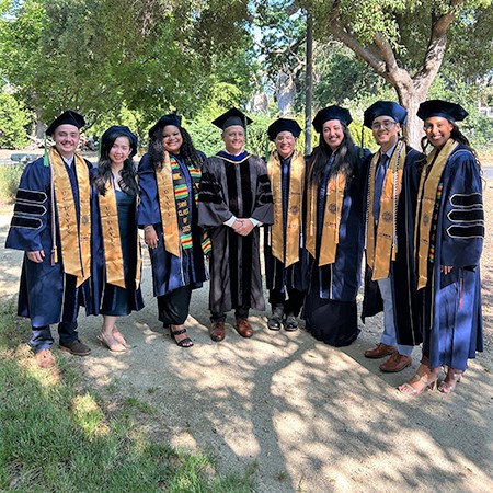A doctor in black gown poses with seven students in graduation garb at the UC Davis medical school&rsquo;s commencement on May 17, 2025.