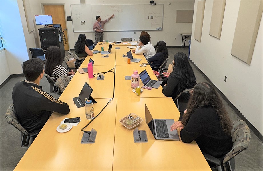 Professor stands at the white board gesturing with left hand before of a classroom where eight students are seated around tables.