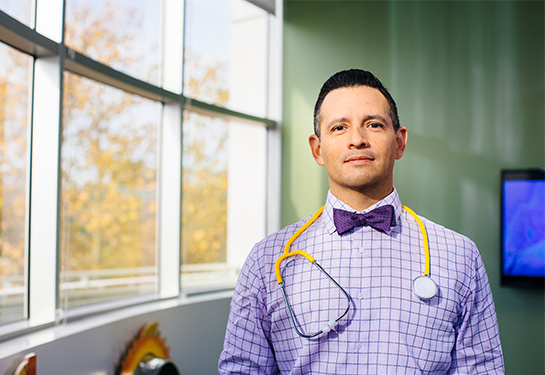 A man with short, dark hair, wearing a purple bow tie and purple shirt, with stethoscope around his neck.