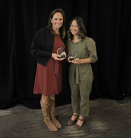 Two women stand next to each other holding glass awards.