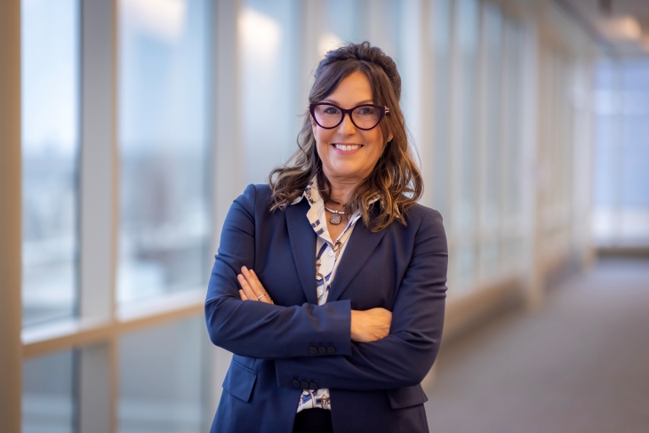 A smiling woman in glasses and a blue suit stands with arms crossed in a hallway with windows.
